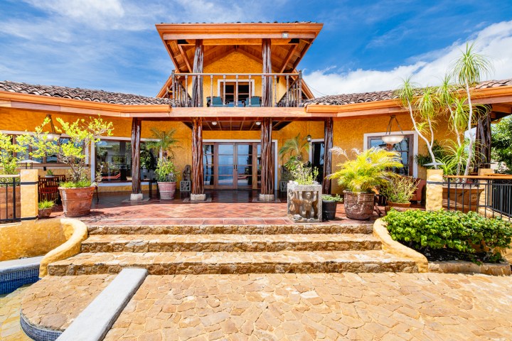 Sunny, tropical villa entrance with plants and a balcony, under a blue sky.