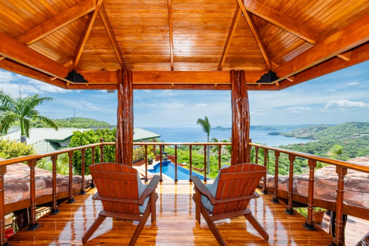 Wooden deck with two chairs overlooking ocean and lush hills under a clear sky.