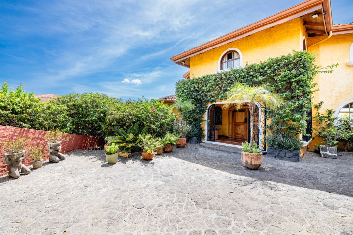 Yellow house with arched doorway, green vines, and potted plants on a stone courtyard under blue sky.