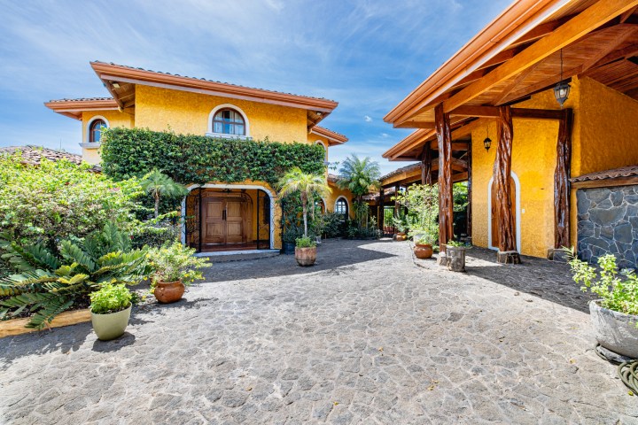 Sunny courtyard with yellow stucco houses, wooden columns, potted plants, and stone pathway.
