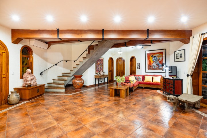 Spacious living room with red sofa, wooden beams, staircase, and art on the walls.