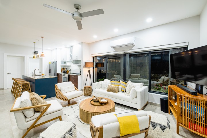 Modern open-plan living room with kitchen, featuring white furniture, wood accents, and a ceiling fan.