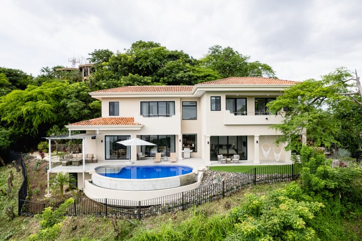 Two-story beige house with red roof, hillside view, pool, and greenery.