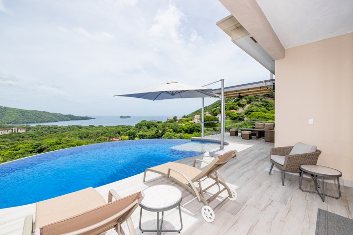 Infinity pool on patio with ocean view, lounge chairs, and umbrella.