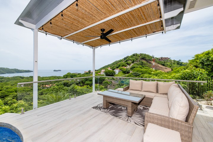 Outdoor patio with cushioned seating, glass railing, and ocean view under a bamboo-covered pergola.