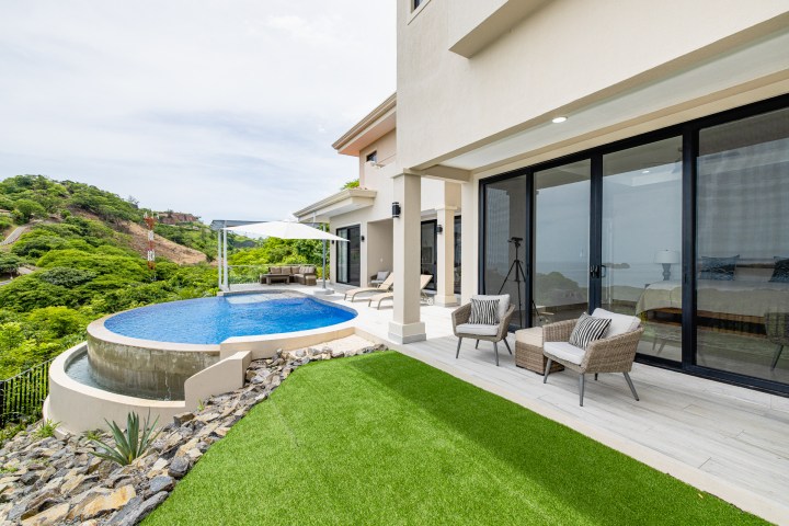 Modern villa with infinity pool, patio chairs, large windows, overlooking lush hills and ocean view.
