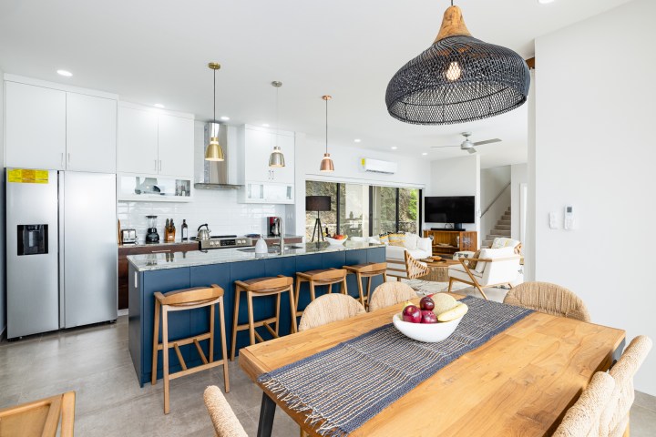 Modern kitchen and living area with wooden dining table, bar stools, and hanging light fixtures.