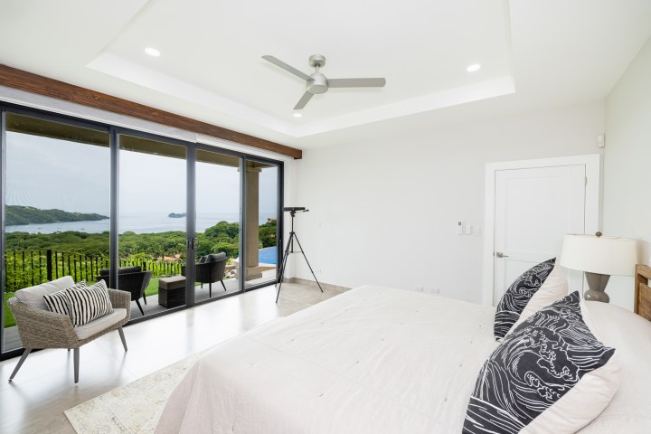 Modern bedroom with view of ocean and greenery through large windows, featuring bed, chairs, and telescope.