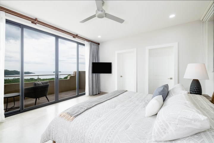 Minimalist bedroom with ocean view, large window, bed, ceiling fan, and balcony with chairs.