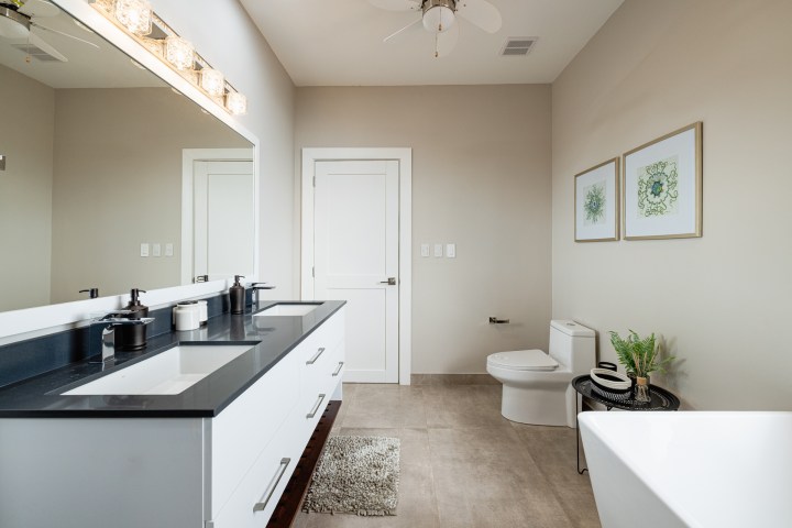 Modern bathroom with double sink, toilet, bathtub, and framed art on beige walls.