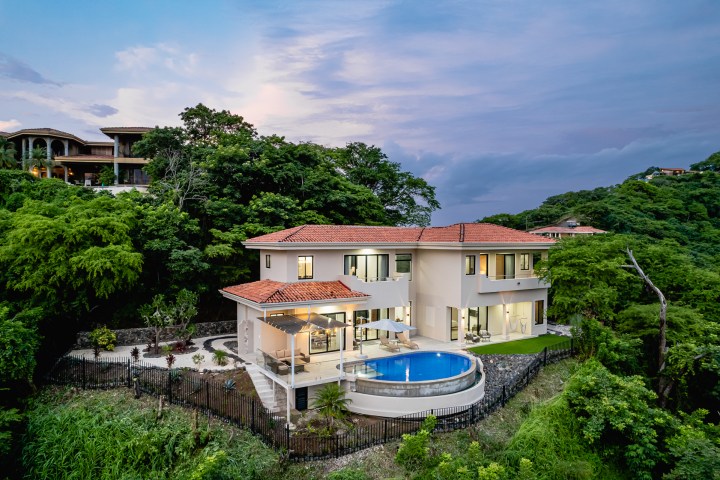 Luxury villa with red roof, surrounded by greenery, featuring a pool and patio at dusk.
