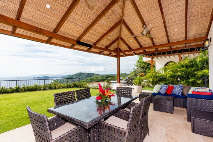 Open-air patio with wicker furniture and ocean view, under a wooden pergola with ceiling fan.