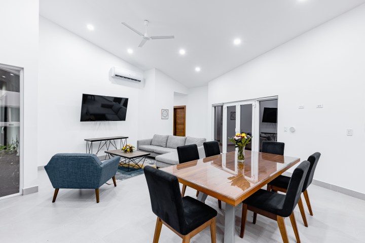 Modern living room with dining area, sofa, TV, and ceiling fan, featuring minimalist decor and bright lighting.