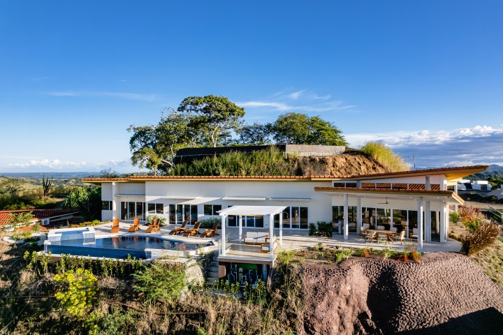 Modern house with rooftop garden, pool, and terrace in a hillside setting under a clear blue sky.