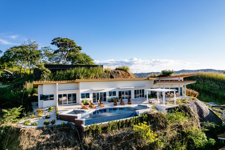 Modern hillside house with pool, patio, and greenery under a clear blue sky.