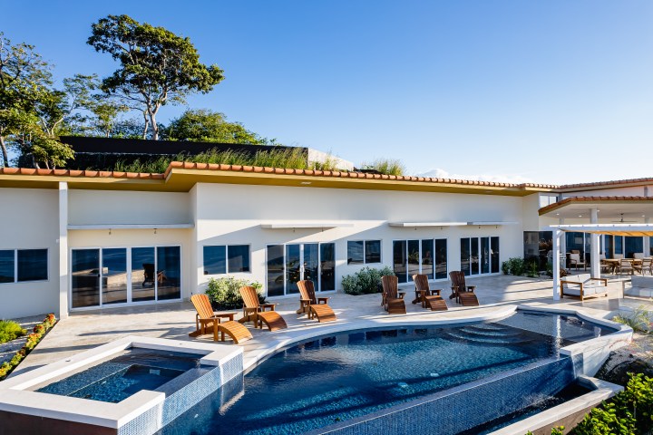 Modern house with large windows, patio, pool, lounge chairs, and trees under a clear blue sky.