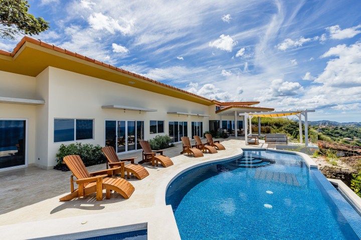 Modern house with pool, wooden loungers, and clear skies in a hillside setting.