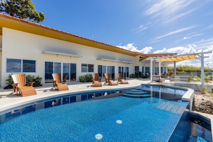 Modern house with patio, wooden chairs, and a pool under a clear blue sky.
