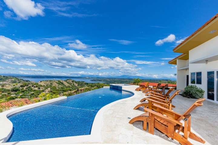 Infinity pool with scenic mountain and ocean view, wooden chairs on deck under clear blue sky.
