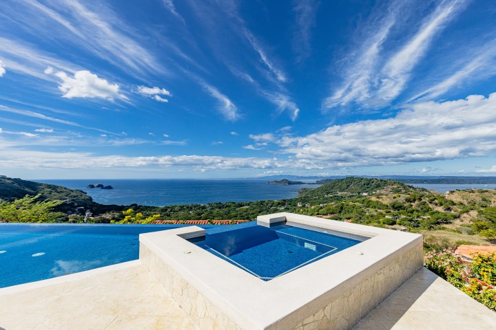 Infinity pool with ocean and hilly landscape view under a clear blue sky.