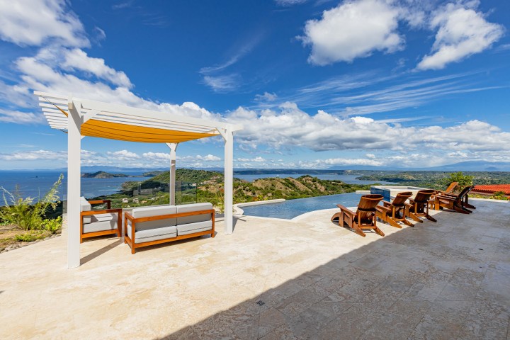 Terrace with pergola, seating, and pool overlooking ocean and hills under a cloudy sky.