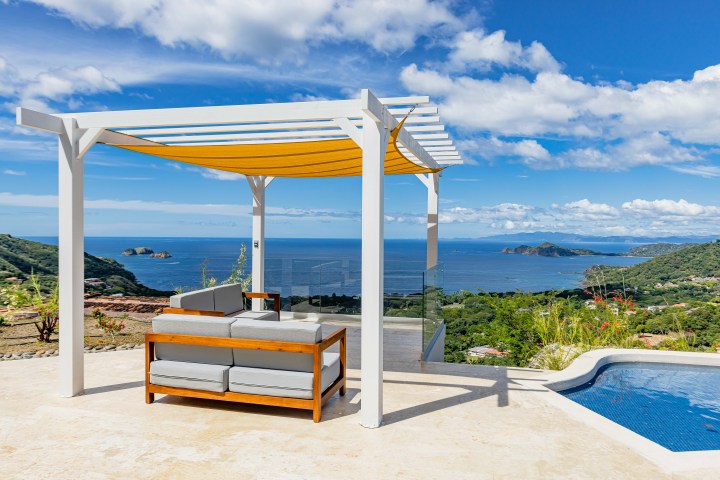 Outdoor lounge with pergola overlooking ocean and hills under blue sky.
