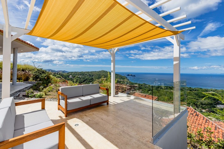 Terrace with seating, canopy, ocean view, and blue sky with clouds.