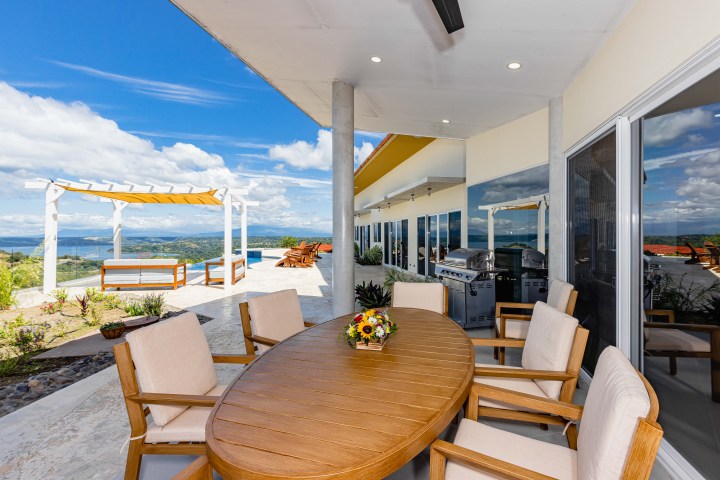 Outdoor patio with wooden table, chairs, pergola, and scenic view of hills and sky.