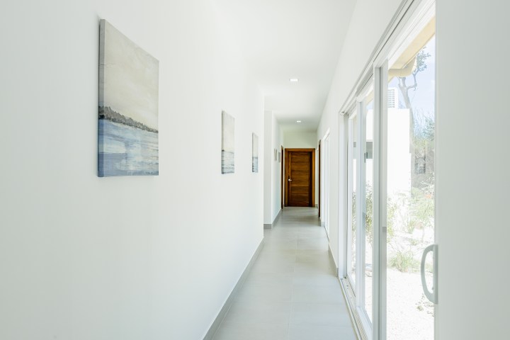 Bright hallway with paintings on left, glass doors on right leading outside, and a wooden door at the end.