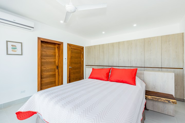 Minimalist bedroom with a white bed and red pillows, wooden doors, and a wall-mounted fan.
