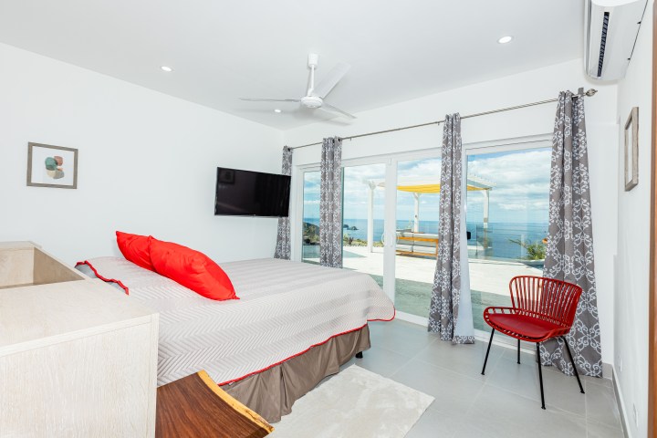 Bright bedroom with red chair, TV, fan, and ocean view through sliding glass doors.