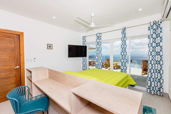 Bright bedroom with sea view, green bed, blue curtains, and wooden furnishings.