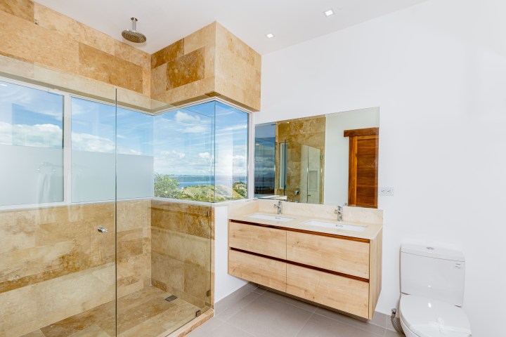 Modern bathroom with beige tiles, glass shower, dual sinks, and ocean view through large window.