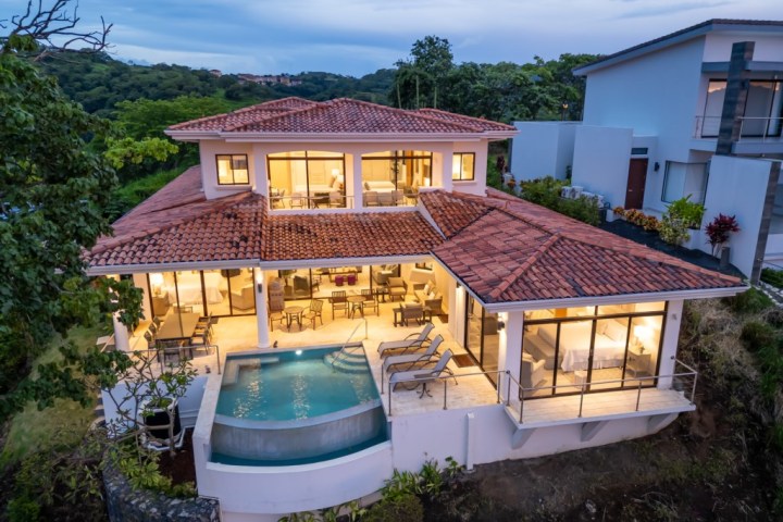 Aerial view of a modern house with red tile roof, lit interior, and a small pool, set among greenery.
