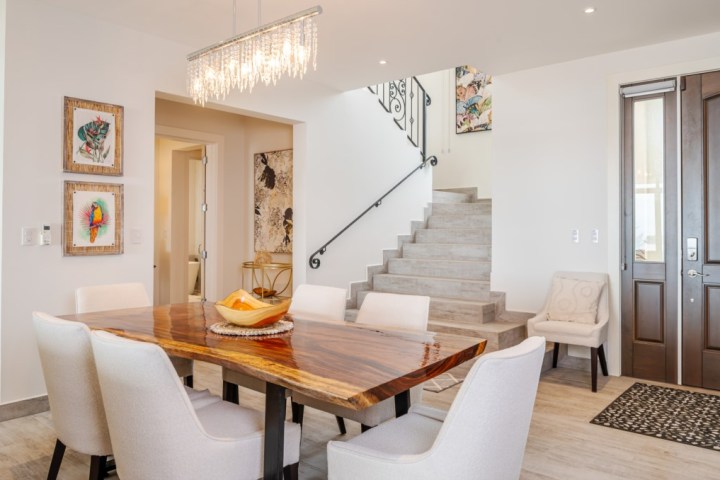 Modern dining room with wooden table, white chairs, and staircase, under a chandelier.