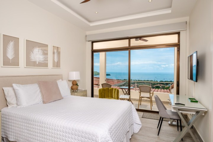 Bedroom with a large bed, ocean view through sliding glass door, desk, and wall-mounted TV.