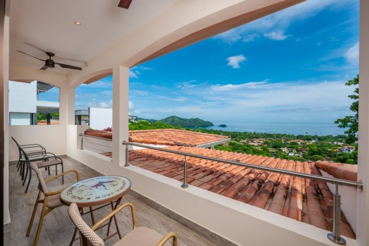 Balcony with chairs overlooks rooftops, lush landscape, and ocean under a clear blue sky.