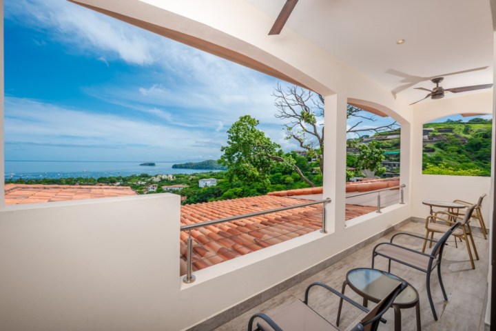 Balcony with chairs overlooking ocean and green hills under a blue sky.