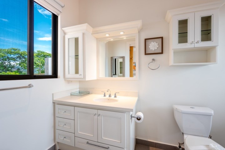 Bright bathroom with white cabinets, mirror, toilet, and window with trees outside.