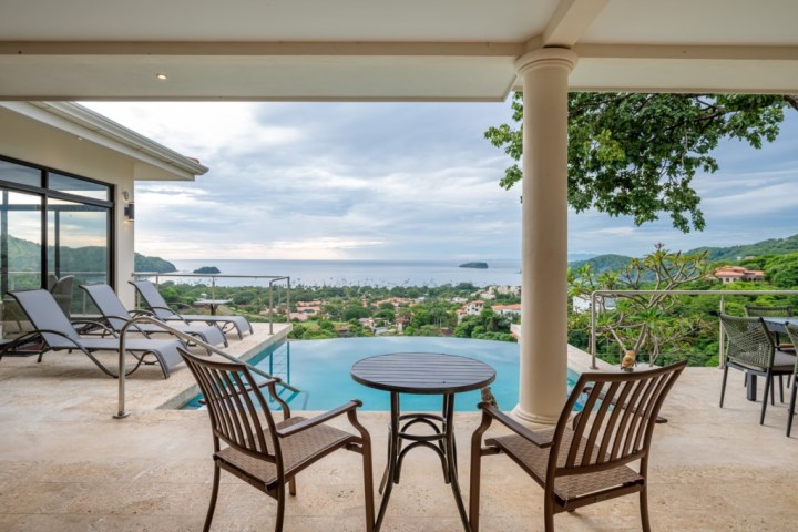 Patio with chairs, infinity pool, and ocean view under a cloudy sky.