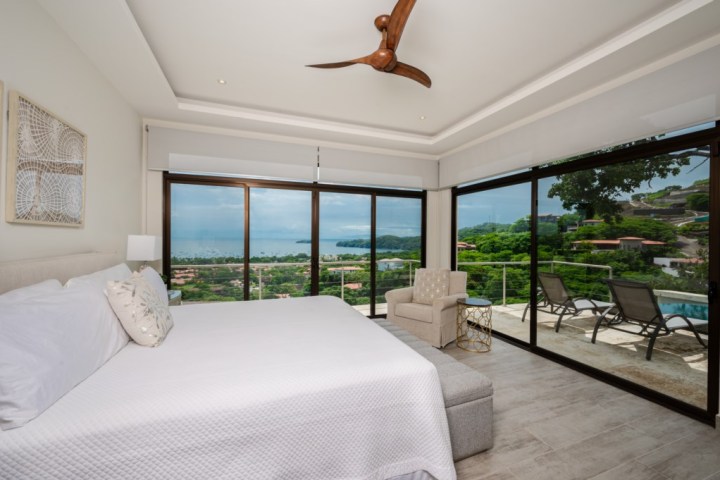 Modern bedroom with ocean view, large windows, white bed, and ceiling fan.