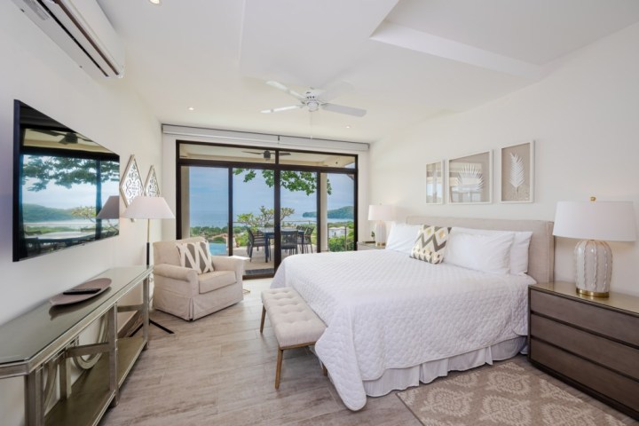 Modern bedroom with ocean view, king bed, TV, beige décor, and large glass doors leading to balcony.