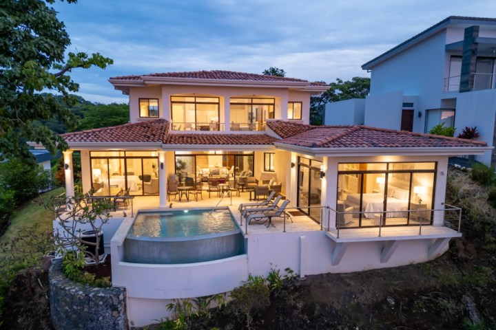 Luxurious modern villa with large windows and an infinity pool on a hillside at dusk.