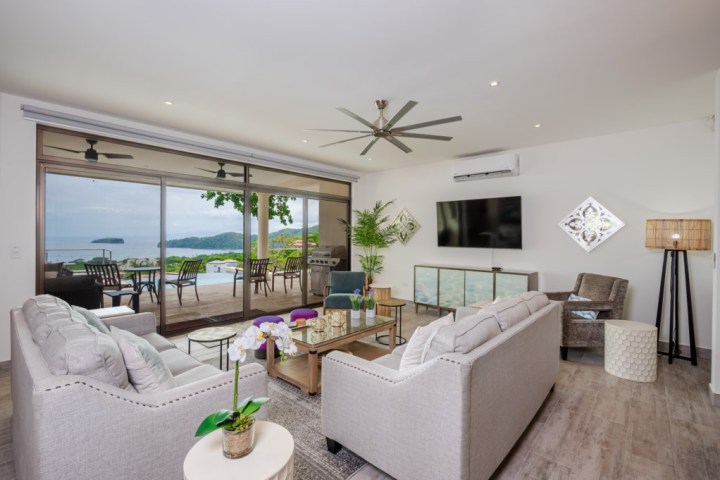 Bright living room with modern furniture, large TV, and sliding doors opening to a patio with ocean view.