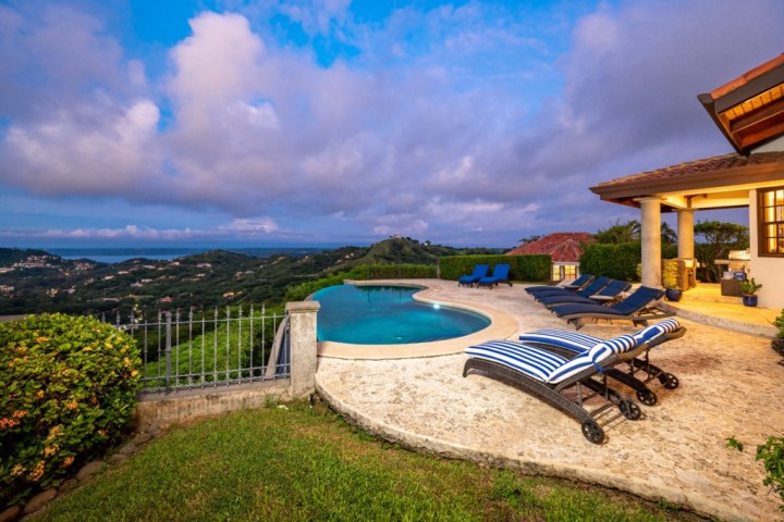 Scenic view of a villa with a pool and lounge chairs overlooking hills and the ocean.