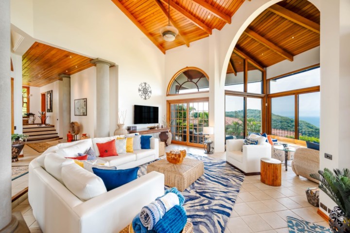 Luxurious living room with white sofas, colorful pillows, large windows, and wooden ceiling.