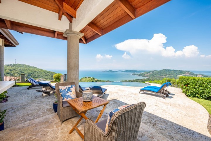 Patio with wicker furniture, infinity pool, and ocean view under a wooden roof.