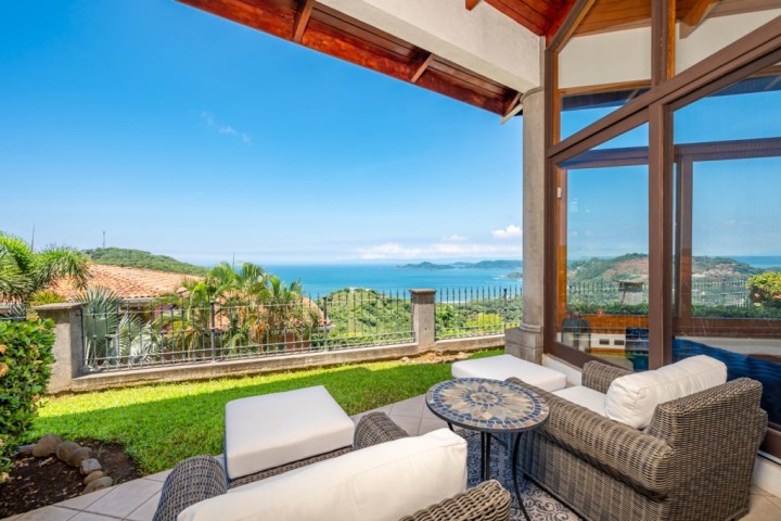 Patio with wicker furniture overlooking tropical landscape and ocean view under clear blue sky.