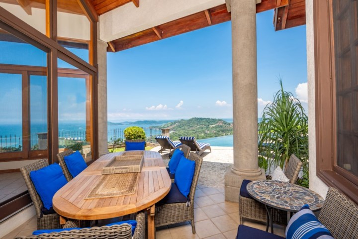 Patio with wicker furniture and blue cushions overlooking ocean and hills.