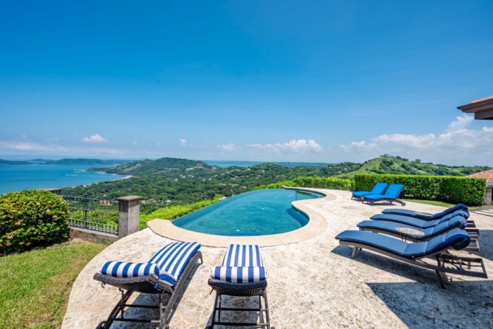 Infinity pool with loungers overlooking ocean and hills under clear blue sky.
